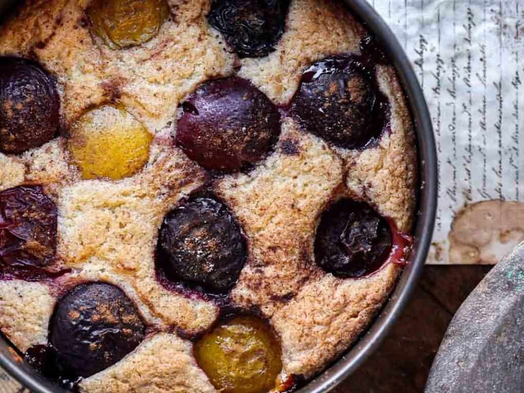A close-up of a rustic vegan plum torte in a round metal baking pan, featuring whole baked plums nestled in golden-brown, slightly textured cake. Handwritten paper is partially visible beneath the pan.